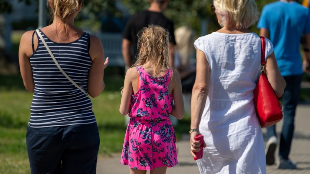 Two women and a young girl in a bright pink floral dress walking along a sunny city street during summer.